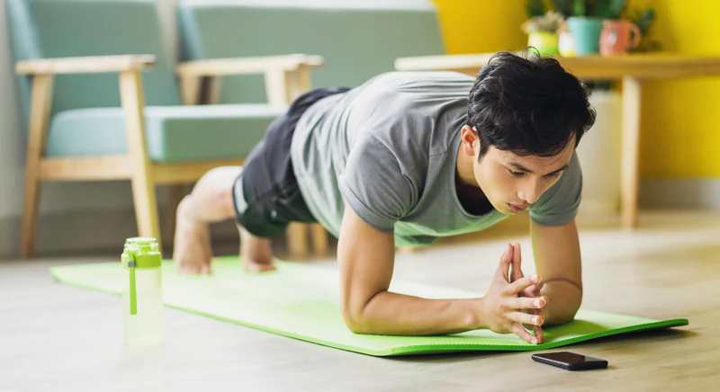 Man doing a plank on a neon green mat at home with a phone in front of him and a water bottle nearby, showcasing an introduction to fitness in a home workout setting.